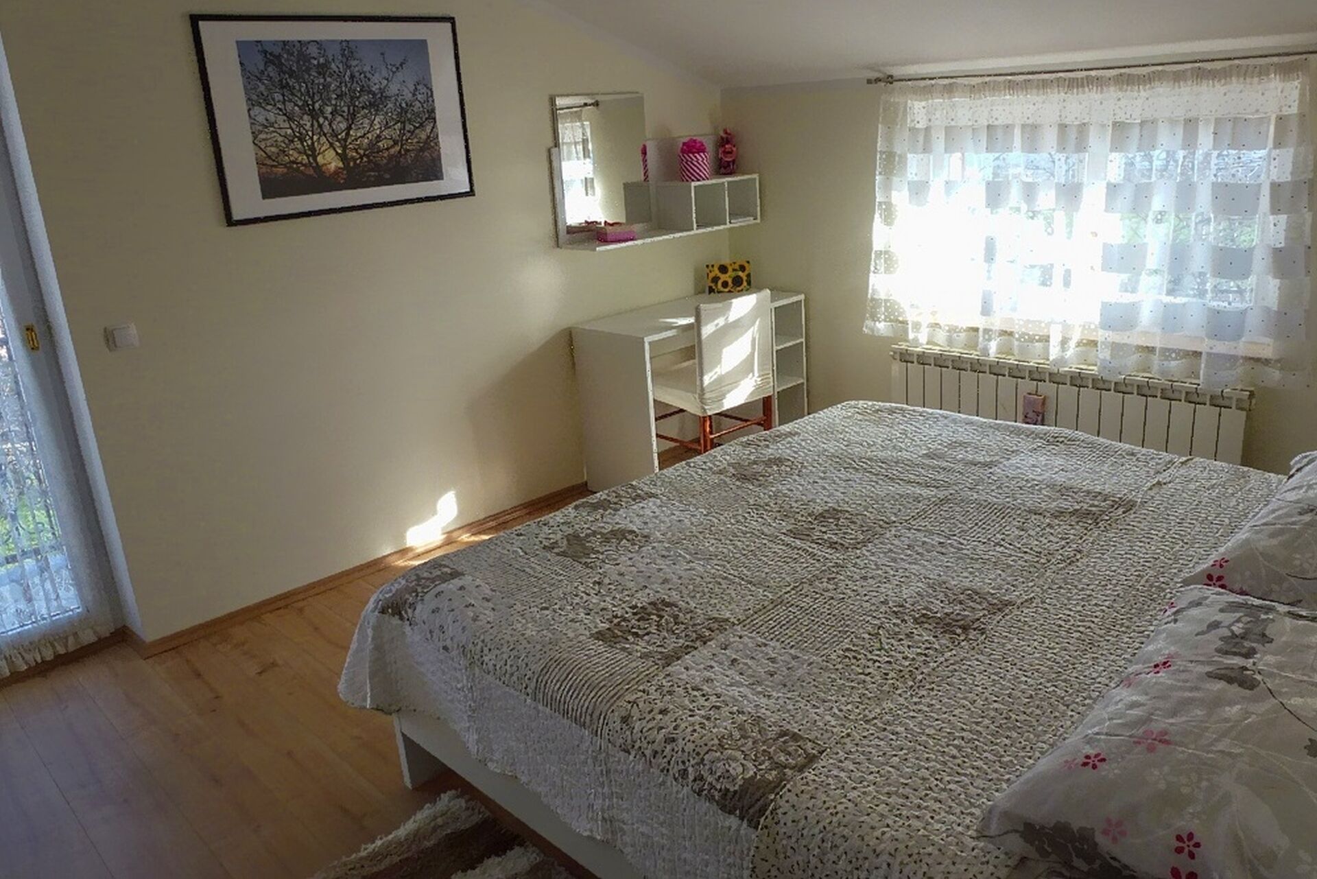 Un dormitorio con una cama grande, un aparador blanco, un cuadro enmarcado en la pared y una ventana con cortinas translúcidas.