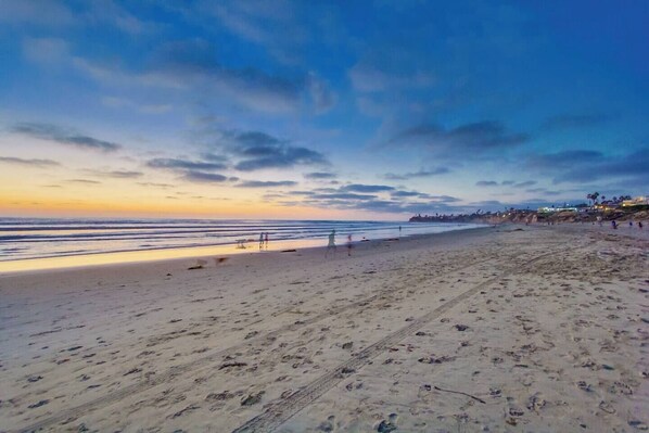 Una playa con una extensa zona arenosa, algunas personas caminando y una costa distante con edificios.