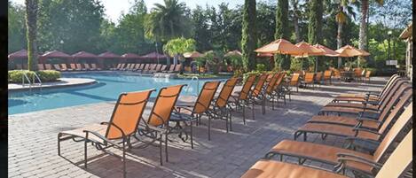 Zona junto a la piscina con sillones de descanso y sombrillas, rodeada de palmeras y un cielo despejado.