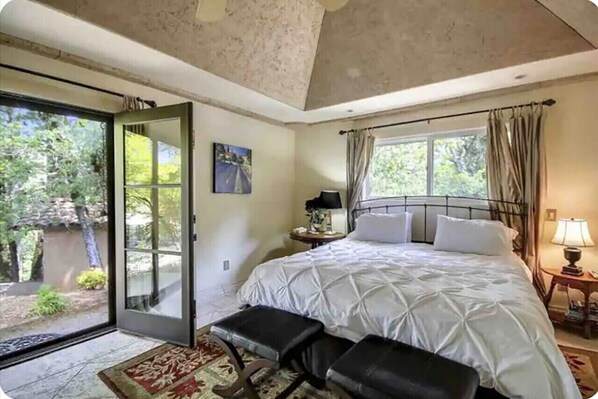 A bedroom with a large bed, a bench, a nightstand, a window with curtains, and a door leading to a balcony.