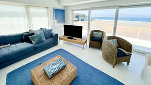 Un salon moderne avec une grande fenêtre donnant sur une plage, un canapé bleu, un fauteuil en osier et une table basse.