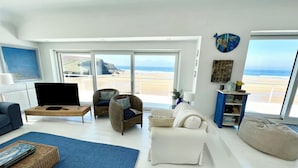 Un salon moderne avec une grande télévision, des fauteuils en osier, un canapé blanc et une vue sur la plage.