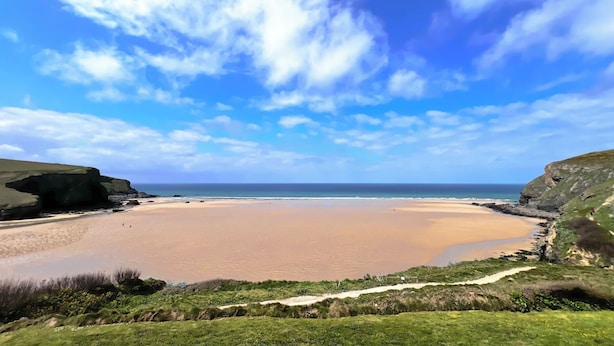 Un paysage côtier avec une plage de sable, des zones herbeuses et des falaises rocheuses, le tout sous un ciel bleu dégagé.