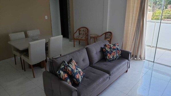 A living room with a grey sofa, two patterned pillows, a dining table with chairs, and a view through a large window.
