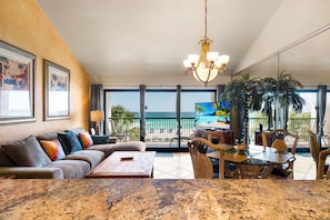 A living room with a sofa, coffee table, and dining table, a chandelier, and a balcony with a view of the ocean.