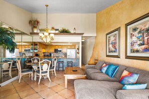 Uma sala de estar com sofá, cadeiras, mesa de centro, área de refeições com mesa e cadeiras, cozinha ao fundo e obras de arte nas paredes.