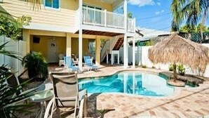 Uma casa de dois andares com piscina, varanda e cobertura de palha.