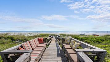 Une promenade en bois mène à un espace de détente sur la plage, avec des coussins à rayures et d’autres de couleur unie.