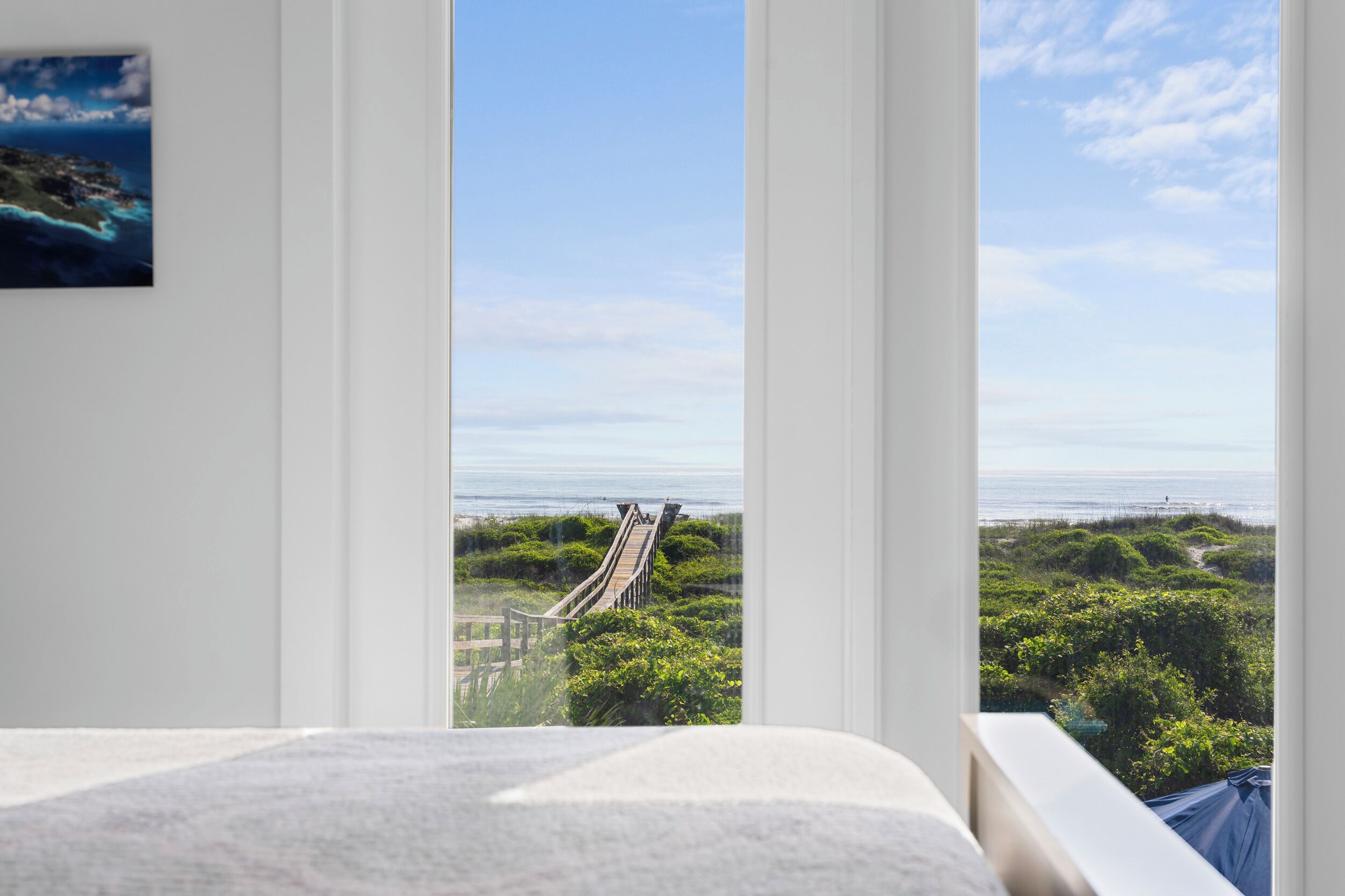 Une chambre dotée d’une grande fenêtre donnant sur un paysage côtier, avec notamment une promenade en bois et de la végétation.