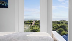 Ein Schlafzimmer mit großem Fenster und Blick auf eine Küstenlandschaft, darunter ein hölzerner Steg und viel Grün.