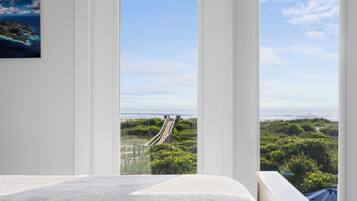 Une chambre dotée d’une grande fenêtre donnant sur un paysage côtier, avec notamment une promenade en bois et de la végétation.
