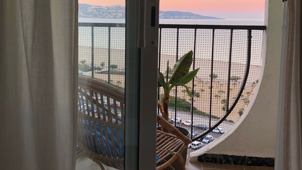 Un balcon donnant sur un plan d’eau et sur des montagnes lointaines, une chaise en osier et une plante en pot.