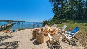 A beach setting with a fire pit, Adirondack chairs, and a dock extending into the water. - Waterfront Retreat w/ Pool, Hot Tub, Dock & Sand Beach. Relax & enjoy the Rivah! (Lancaster)