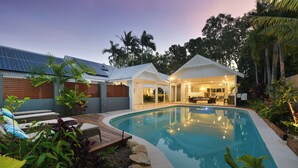 A swimming pool with a wooden deck, a covered outdoor seating area, and a building with a white facade.