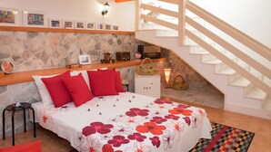 Una camera da letto con parete in pietra, un letto con cuscini rossi, una mensola in legno con quadri incorniciati, una cassettiera bianca e una scala che conduce a un piano superiore.