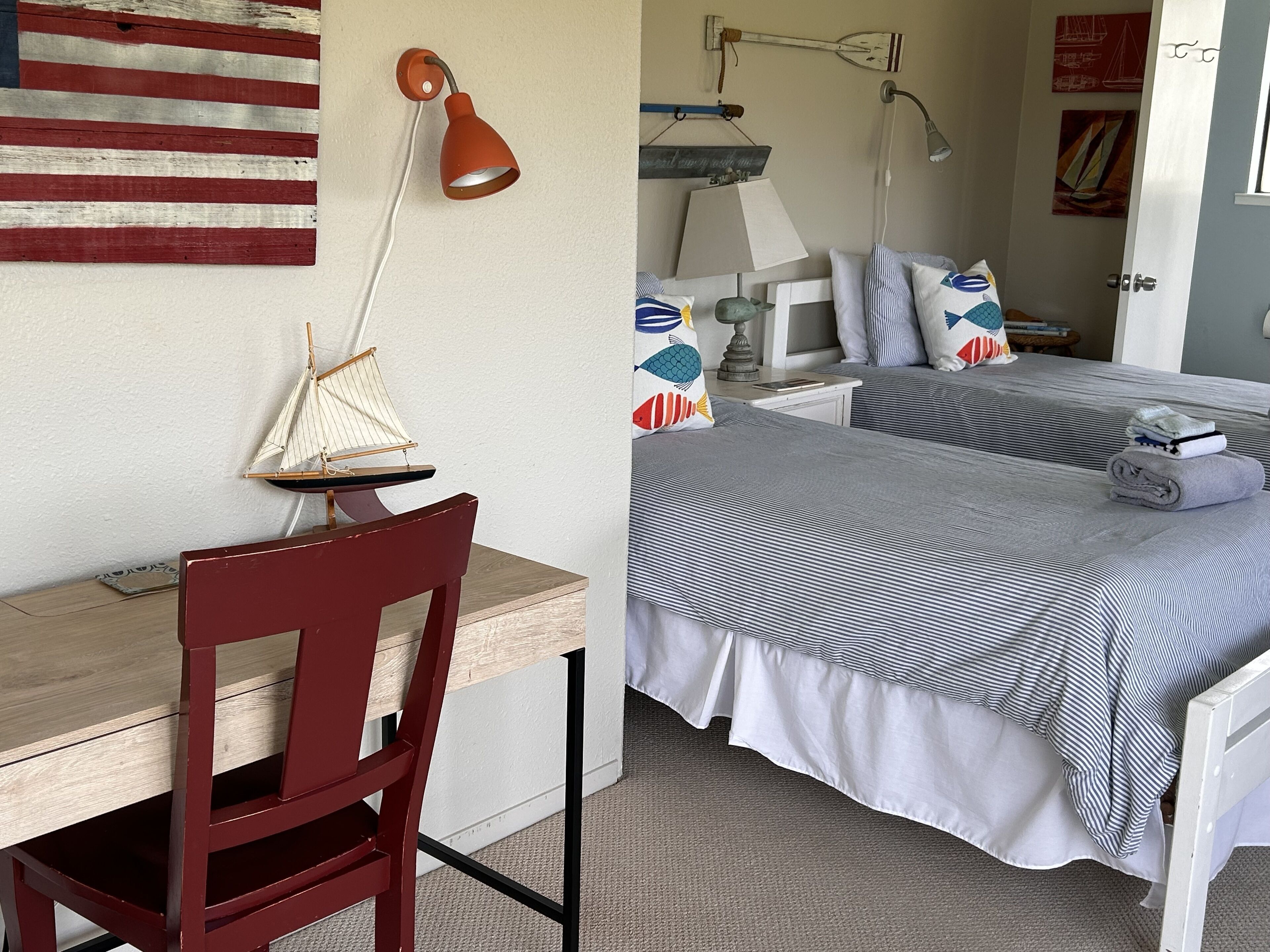 A room with a desk, two beds with striped bedding, a red chair, a wall-mounted lamp, and a small sailboat decoration.