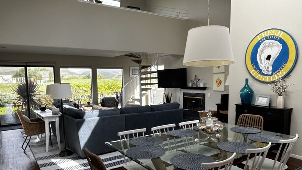 Un salon moderne avec une table à manger en verre, des chaises blanches, un canapé bleu et une grande fenêtre donnant sur un paysage verdoyant.