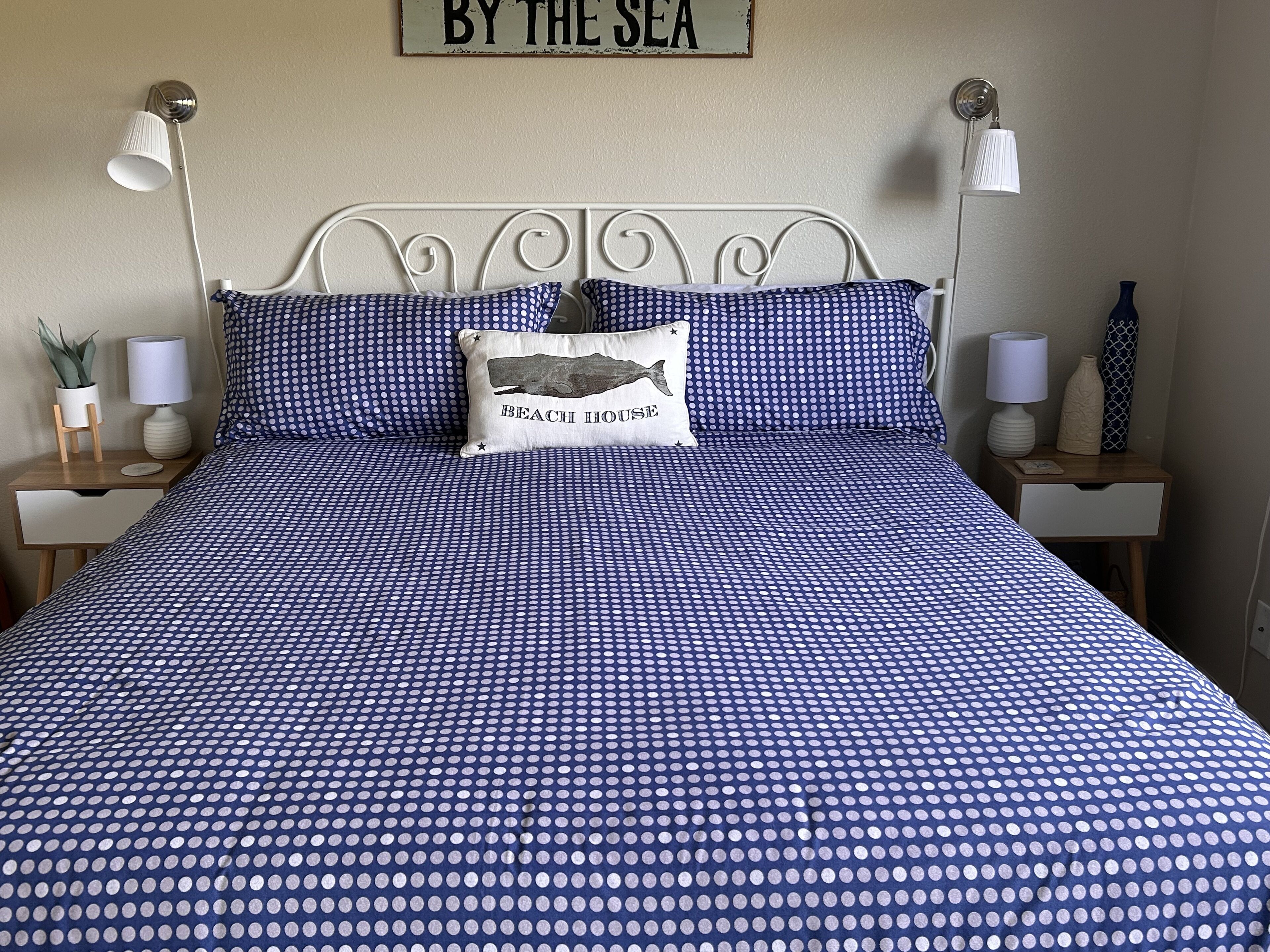 Un lit avec une couvre-lit à pois bleus, un oreiller orné d’un motif de maison de plage et un panneau mural sur lequel on peut lire : « AU BORD DE LA MER ».