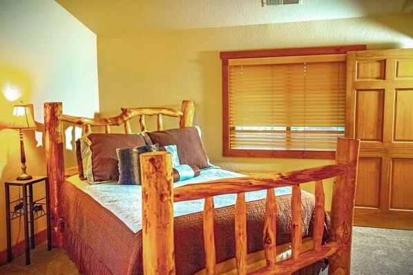 A wooden-framed bed with a headboard, a bedside table with a lamp, and a window with blinds.