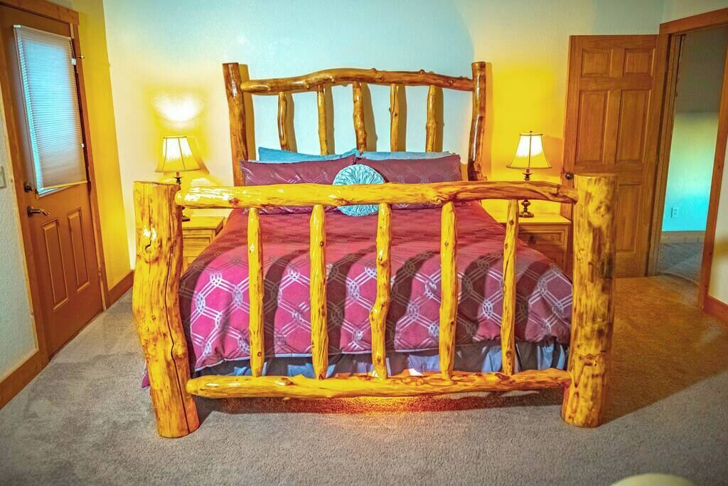 Un lit en bois rustique au design original, doté d’une tête de lit et d’un pied de lit fabriqués à partir de troncs d’arbres. Deux tables de chevet avec des lampes sont placées de chaque côté du lit.