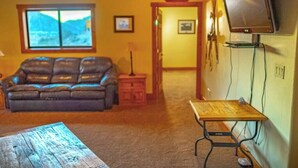 A living room with a leather sofa, a wooden coffee table, a television mounted on the wall, and a window with a view of mountains. - River Ranch (Canon City)