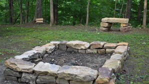 A stone fire pit surrounded by a forested area with a bench in the background.