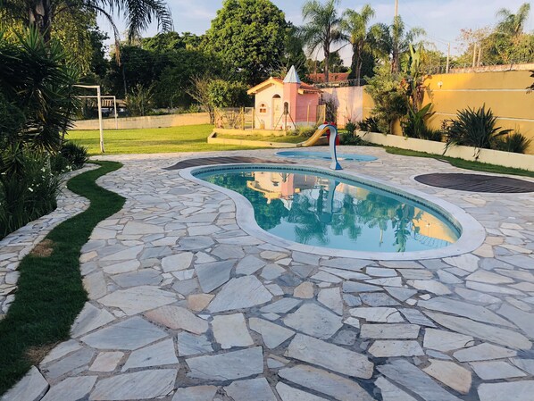 Piscina amb rajoles de pedra i zona verda amb un parc infantil.