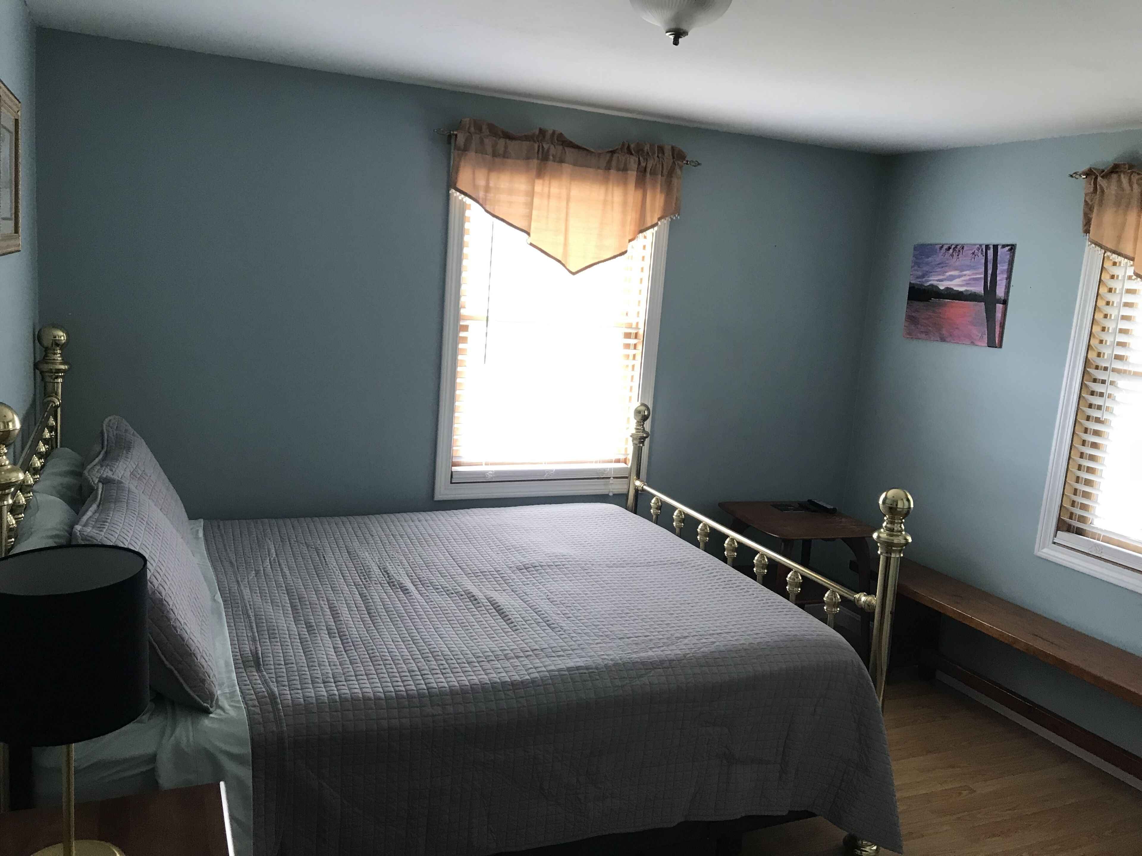 A bedroom with a bed, a nightstand, a lamp, a window with curtains, a painting, and a bench.