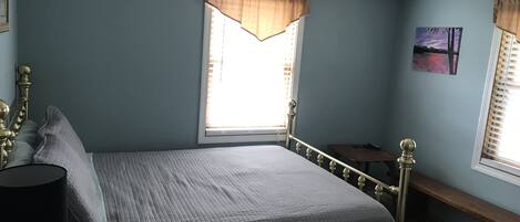 A bedroom with a bed, a nightstand, a lamp, a window with curtains, a painting, and a bench.