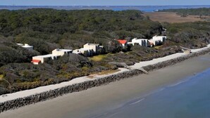 Uma área costeira com vários edifícios brancos e telhados vermelhos, rodeada por vegetação densa e uma praia arenosa.