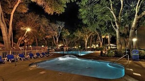 Uma área de piscina à noite, com espreguiçadeiras e uma pequena ponte sobre a piscina.