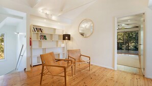A room with wooden flooring, two wooden chairs, a bookshelf, a round mirror, and a door leading to an outdoor area. - Eucalyptus Beach House by HolidayCo. (KILLCARE)