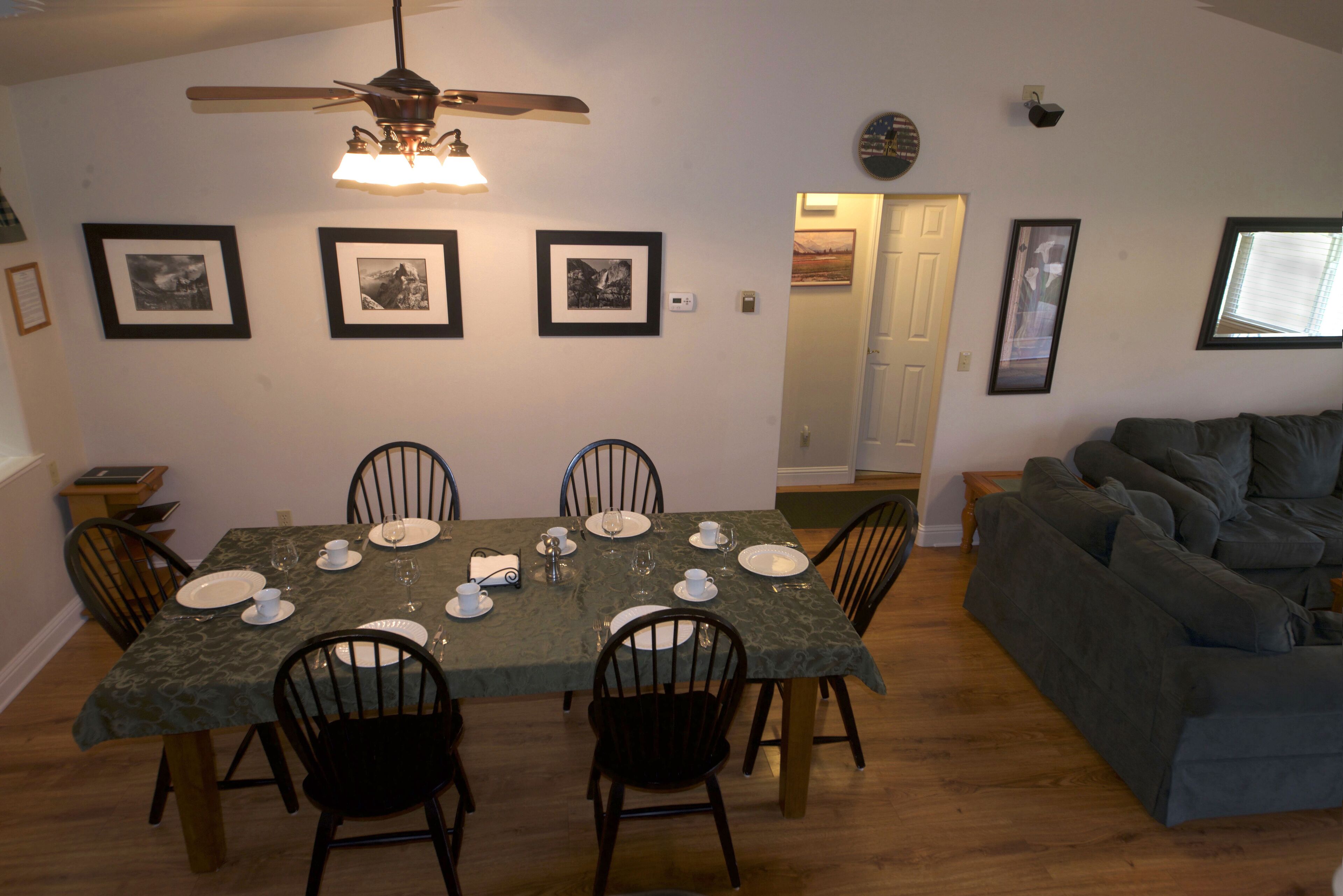 Un coin repas avec une table dressée pour six personnes, entourée de chaises, un ventilateur de plafond et des tableaux encadrés accrochés aux murs.