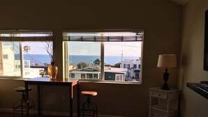 Quarto com uma janela ampla que oferece vista para o mar e edifícios. Há uma mesa com dois banquinhos, um abajur e um pequeno armário.