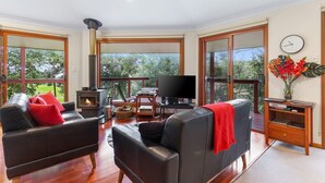 A spacious living room with a fireplace, leather sofas, a television, and large windows offering a view of greenery.