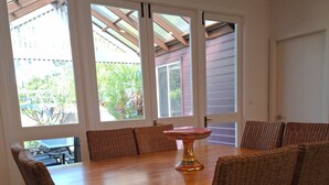 A dining area with a wooden table and wicker chairs, large windows with a view of greenery, and a ceiling fan.