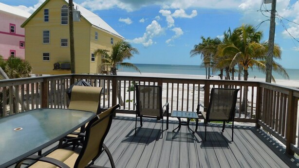 A wooden deck with a table and chairs, overlooking a beach with palm trees and a view of the ocean. - Spectacular Beach Cottage, Pet Friendly, Gulf Views, Great for Families (Fort Myers Beach)