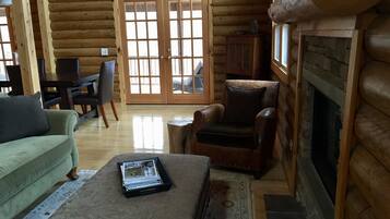 A log cabin interior with a green sofa, a leather armchair, a fireplace, and a dining area with a table and chairs.