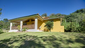 Une maison jaune avec un porche, entourée de verdure et d’arbres.