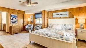 Une chambre avec un mur lambrissé en bois, un lit recouvert d’une couvre-lit à motifs, un ventilateur de plafond et une table de chevet avec une lampe.