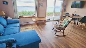 En stue med en blå sofa, en gyngestol, et træbord og havudsigt gennem store vinduer.