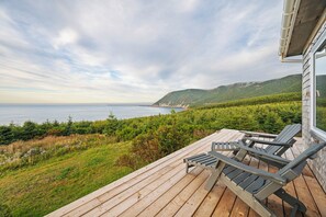 A wooden deck with two Adirondack chairs overlooking a coastal landscape with mountains in the distance. - Peaceful Cape Breton cottage with breathtaking ocean and mountain views! (Victoria, Subd. A)