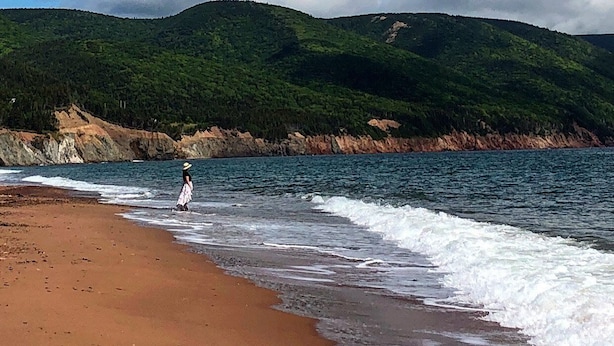 En strand med en person, der går langs kysten, stenet kystlinje og grønne bakker i baggrunden.
