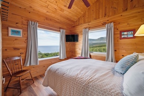 A wooden cabin room with a bed, a chair, and two windows offering a view of the sea. - Peaceful Cape Breton cottage with breathtaking ocean and mountain views! (Victoria, Subd. A)