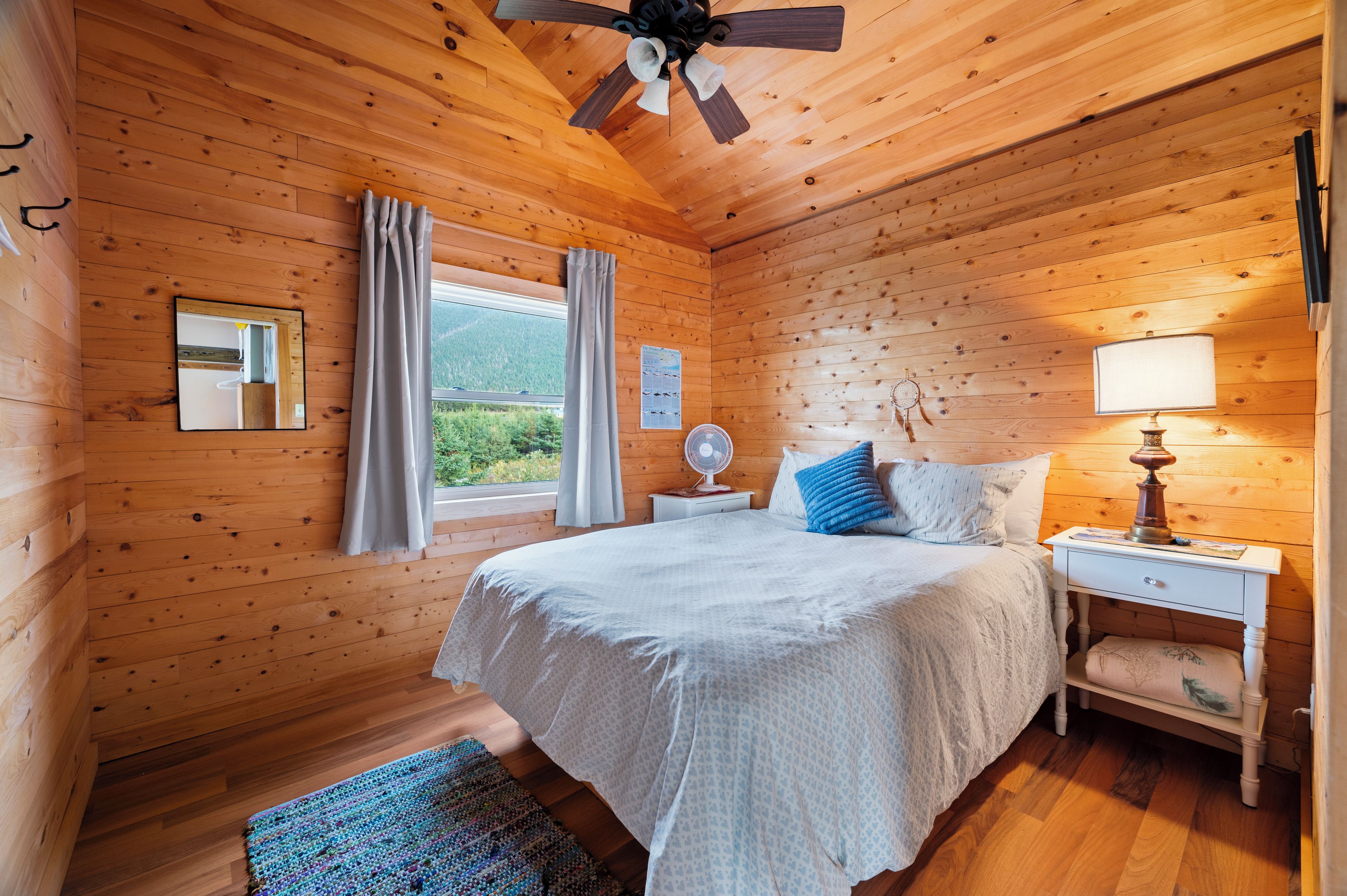 Une chambre dans une cabane en bois, avec un lit, une table de chevet, une lampe et une fenêtre avec des rideaux.