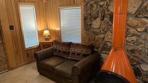 A cozy room with a stone wall, wooden paneled walls, a brown leather sofa, and a large orange chimney. - Mountain-side Cabin with hot tub! Close To Town And Skiing on 1.1 acres.  (Ruidoso)