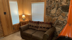 A cozy room with a brown leather sofa, a stone wall, and a window with blinds. - Mountain-side Cabin with hot tub! Close To Town And Skiing on 1.1 acres.  (Ruidoso)