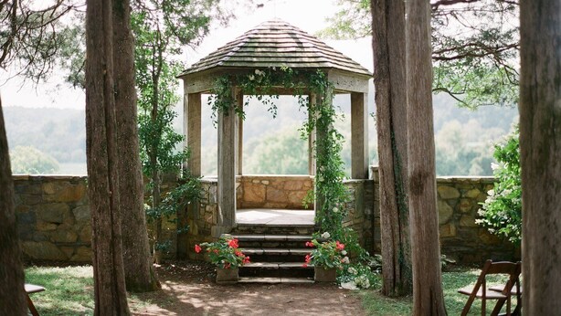 A gazebo with a stone bench, surrounded by trees and a stone wall, overlooking a scenic landscape.