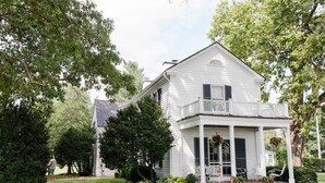 A white two-story house with a balcony, surrounded by lush greenery and trees.