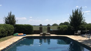 A rectangular swimming pool with a stone border, surrounded by a hedge and trees, with a clear sky above.